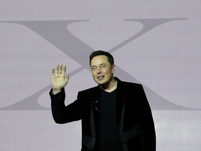 Elon Musk in a black suit waving with his right hand and a large 'X' on a white wall behind him