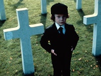 A boy standing in a graveyard while wearing a black suit in the horror movie 'The Omen' from the Inv...
