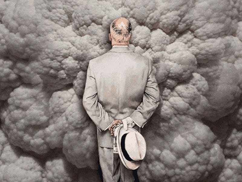 A man in a grey suit with his hands behind his back walking into smoke caused by an explosion
