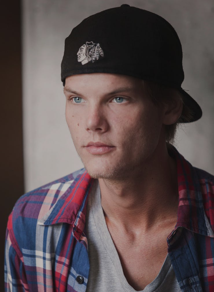 A close-up portrait of Avicii in a white T-shirt, blue-red check shirt and a black cap
