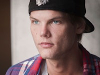 A close-up portrait of Avicii in a white T-shirt, blue-red check shirt and a black cap