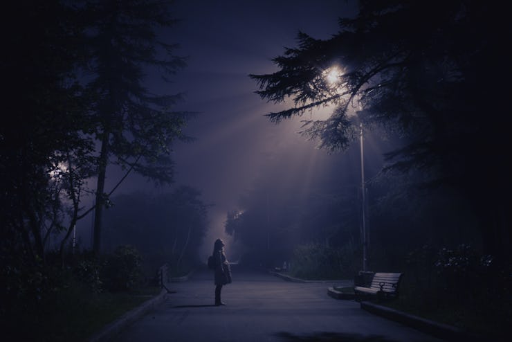 A woman standing on a park path with a UFO's light shining through the trees in front of her