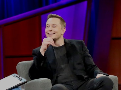 Elon Musk smiling in a black shirt and black blazer during an interview 