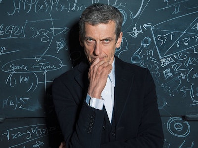 Peter Capaldi in a white shirt and black blazer in front of a blackboard
