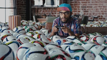 A man focused on playing video games on PlayStation in a room filled with soccer balls