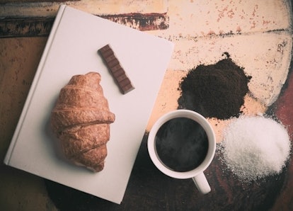 A plate with a croissant and chocolate, next to a cup of coffee that is surrounded with coffee and s...