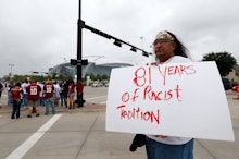 A man standing in a parking lot holding a white poster with red text '81 years of racist tradition'