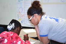 A teacher with curly brunette hair assisting a student in a classroom