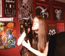 A metalhead posing with his black cat in a room with walls with posters