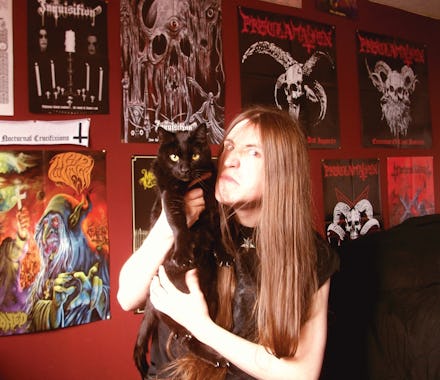 A metalhead posing with his black cat in a room with walls with posters