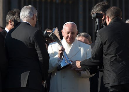 Pope Francis in a white coat, surrounded by journalists photographing him, while he is holding a sil...