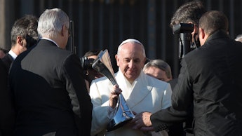 Pope Francis in a white coat, surrounded by journalists photographing him, while he is holding a sil...