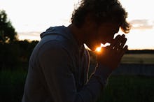 A man standing outside with his hands in a praying gesture position with the sunset in the backgroun...