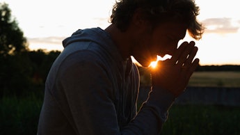 A man standing outside with his hands in a praying gesture position with the sunset in the backgroun...