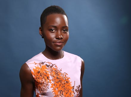 Lupita Nyong'o posing, with her hands behind her back, and a dark- blue backdrop, wearing a white sl...