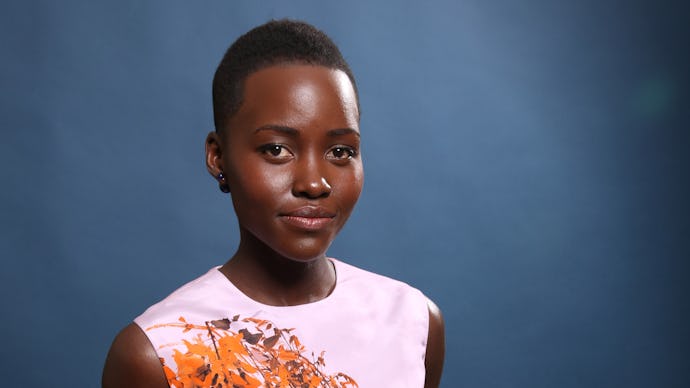 Lupita Nyong'o posing, with her hands behind her back, and a dark- blue backdrop, wearing a white sl...