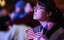 A girl sitting down, holding a small American flag, looking upwards