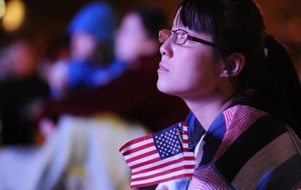 A girl sitting down, holding a small American flag, looking upwards