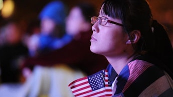 A girl sitting down, holding a small American flag, looking upwards