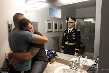 Two men hug in a bathroom with a reflection of a soldier in uniform behind them