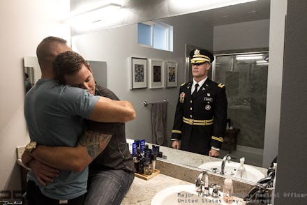 Two men hug in a bathroom with a reflection of a soldier in uniform behind them