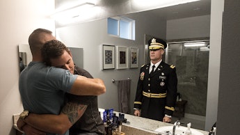 Two men hug in a bathroom with a reflection of a soldier in uniform behind them