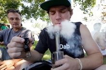 A guy sitting on the grass, smoking marijuana from a bong, and another guy, on his right, watching h...