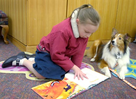 A girl sitting on a floor while reading a book and a dog sitting next to her and looking at her