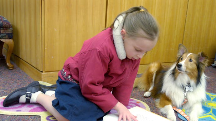 A girl sitting on a floor while reading a book and a dog sitting next to her and looking at her