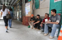 Construction workers sitting on a sidewalk, enjoying their drinks, observing the pedestrians that ar...