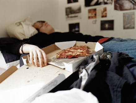An adult slob lying in bed with their hand in a half empty pizza box