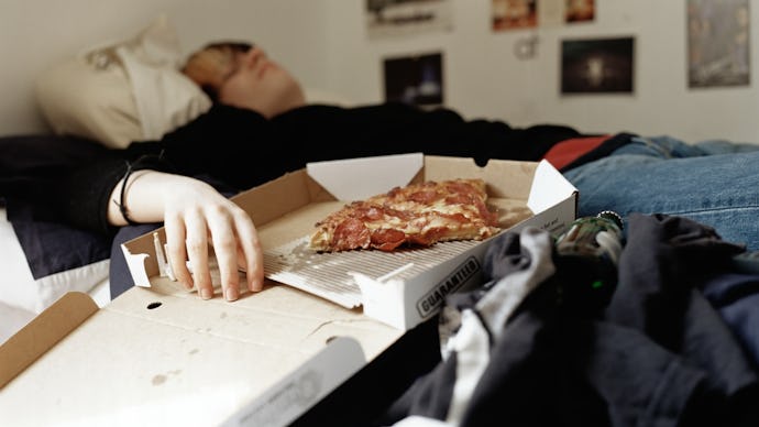 An adult slob lying in bed with their hand in a half empty pizza box