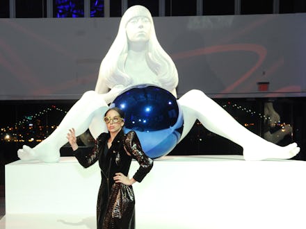 Lady gaga in front of a large white statue of a woman holding a blue ball between her legs