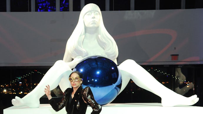 Lady gaga in front of a large white statue of a woman holding a blue ball between her legs