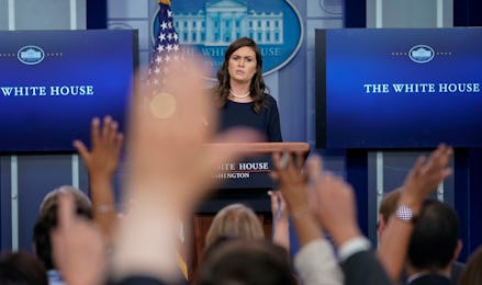 White house spokesperson standing in front of a podium while journalists raise their hands to ask qu...