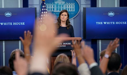 White house spokesperson standing in front of a podium while journalists raise their hands to ask qu...