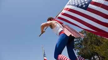 A man riding a unicycle in a white tank, blue pants and striped red and white socks, with an America...