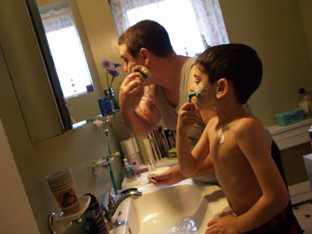 A man shaving his face in the bathroom while his son stands next to him, mimicking his behavior