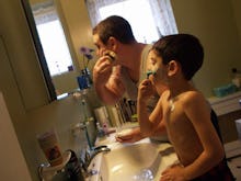 A man shaving his face in the bathroom while his son stands next to him, mimicking his behavior