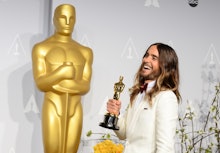 Jared Leto in a white suite, smiling, with an Oscar in his hand, in front of a huge Oscar statue. 