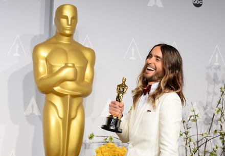 Jared Leto in a white suite, smiling, with an Oscar in his hand, in front of a huge Oscar statue.
