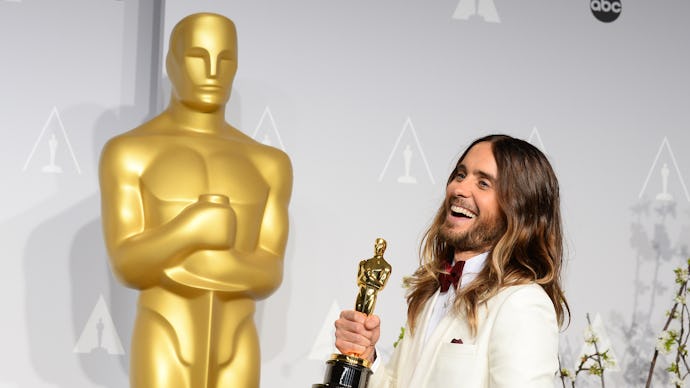 Jared Leto in a white suite, smiling, with an Oscar in his hand, in front of a huge Oscar statue. 