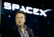 Elon Musk Speaking in a black jacket and a black and white shirt and the SPACEX logo behind him