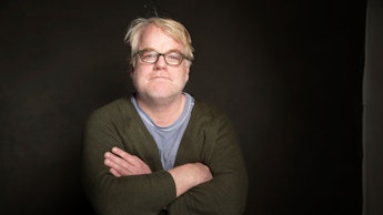 Philip Seymour Hoffman with his arms crossed, in front of a dark backdrop, with a slight grin on his...