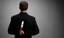A man in a black suit facing away from the camera holding a large knife hidden behind his back