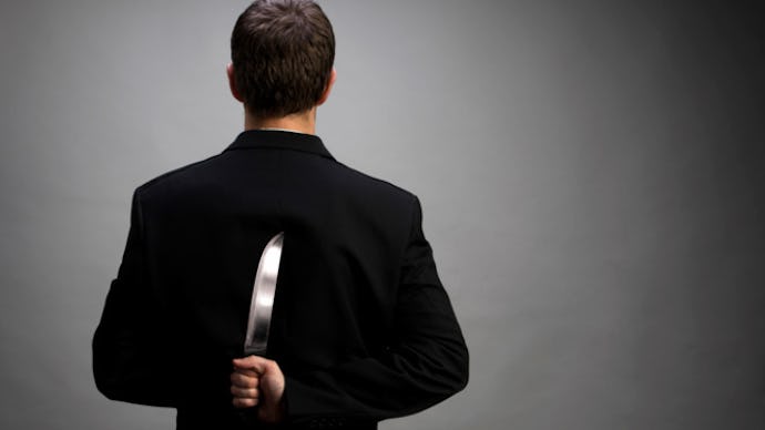 A man in a black suit facing away from the camera holding a large knife hidden behind his back