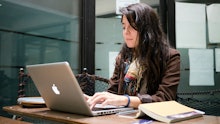 A college student typing on her laptop in the classroom with a book beside her.