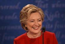 Hillary Clinton in a red suit laughing with a microphone in front of her