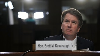 Brett Kavanaugh sitting down behind a plaque with his name on it, lights shine behind him
