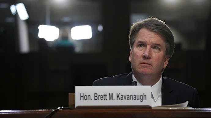 Brett Kavanaugh sitting down behind a plaque with his name on it, lights shine behind him